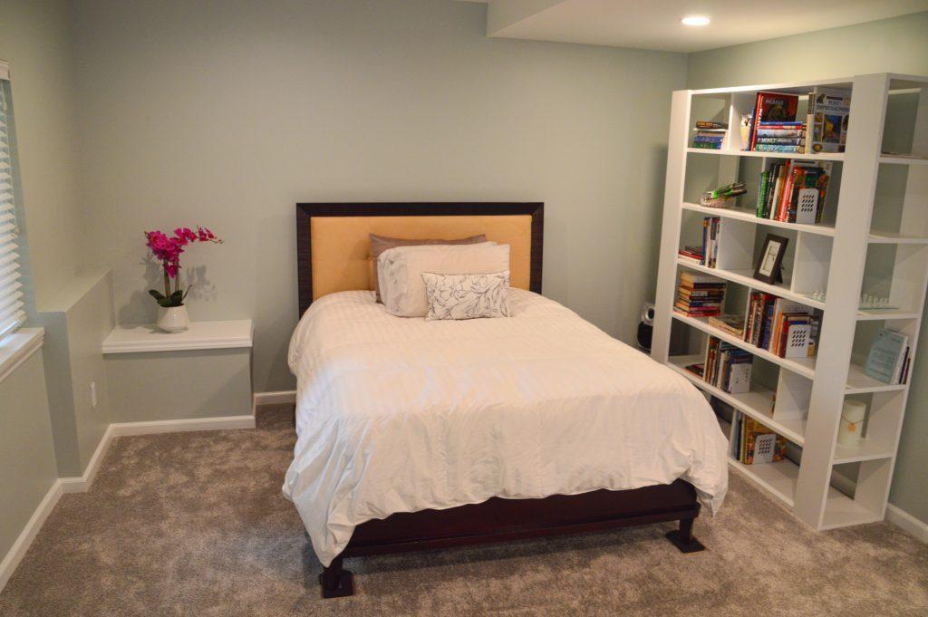 Close up of finished basement guest bed area displaying the sump pump cover, bed and shelving area with a collection of books,