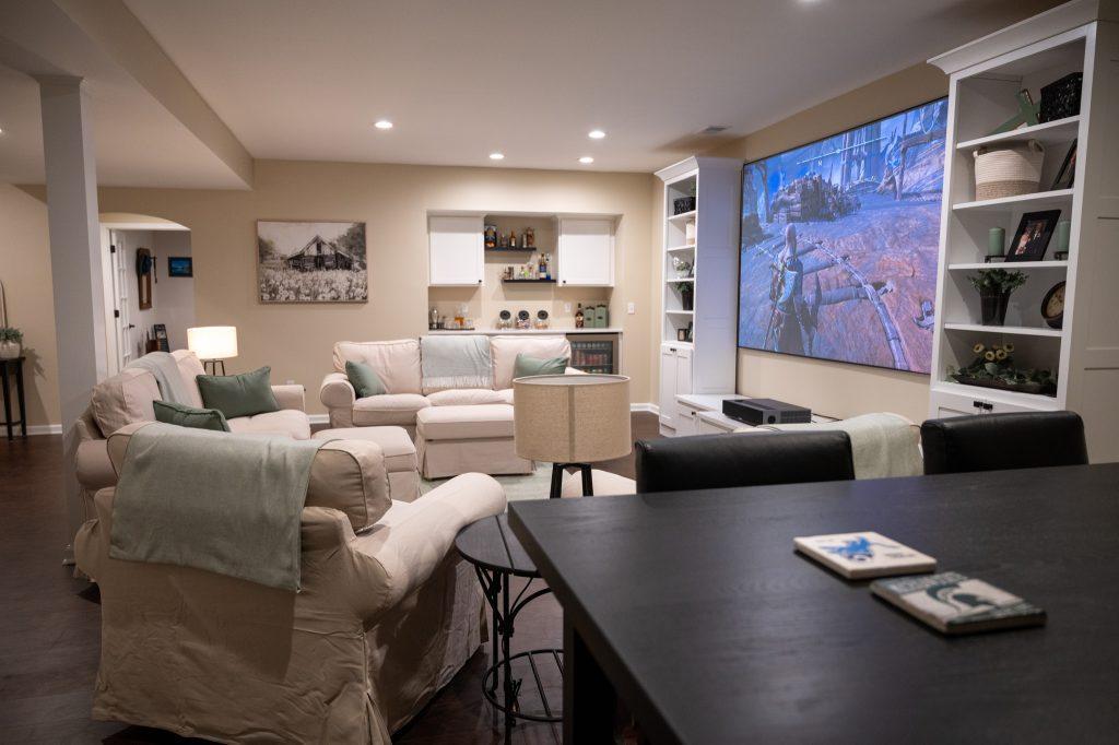 Long view of a finished basement showing a table for sitting, projection screen for movies and video games along with a snack bar in the back.