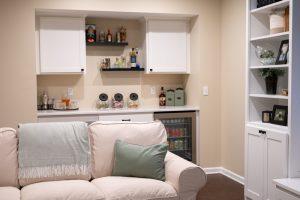 Finished basement living room area with dry bar in the background with spirits and snacks for watching movies along with a peek at the shelving unit.